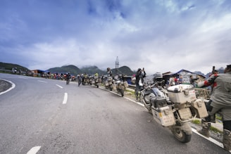 A group of motorcyclists is parked along the side of a winding mountain road. The riders, wearing helmets and gear, are gathered around and some are taking photos, possibly capturing the scenic view. The background features mist-covered mountains and a cloudy sky, with a few makeshift structures or stalls visible along the road.