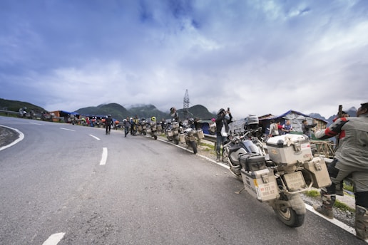 A group of motorcyclists is parked along the side of a winding mountain road. The riders, wearing helmets and gear, are gathered around and some are taking photos, possibly capturing the scenic view. The background features mist-covered mountains and a cloudy sky, with a few makeshift structures or stalls visible along the road.