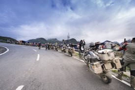A group of motorcyclists is parked along the side of a winding mountain road. The riders, wearing helmets and gear, are gathered around and some are taking photos, possibly capturing the scenic view. The background features mist-covered mountains and a cloudy sky, with a few makeshift structures or stalls visible along the road.