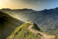 A breathtaking mountain landscape with a group of people standing on a grassy peak, surrounded by rolling hills and layers of distant mountains under a hazy sky.