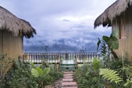 Two thatched-roof huts flank a lush garden path leading to a wooden deck. The background showcases a stunning mountain range shrouded in mist and clouds.
