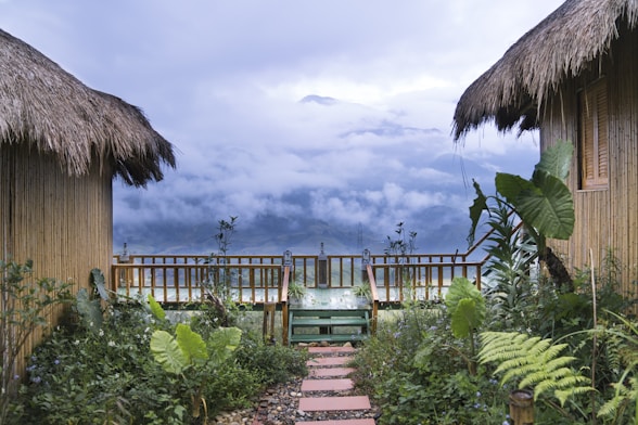 Two thatched-roof huts flank a lush garden path leading to a wooden deck. The background showcases a stunning mountain range shrouded in mist and clouds.