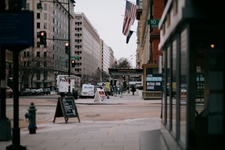 An urban street scene with tall buildings lining both sides. A FedEx truck and other vehicles are on the road. People are walking on the sidewalks, and there are signs advertising retail space for lease. Flags hang from the buildings, and the atmosphere is overcast and calm.