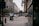 An urban street scene with tall buildings lining both sides. A FedEx truck and other vehicles are on the road. People are walking on the sidewalks, and there are signs advertising retail space for lease. Flags hang from the buildings, and the atmosphere is overcast and calm.