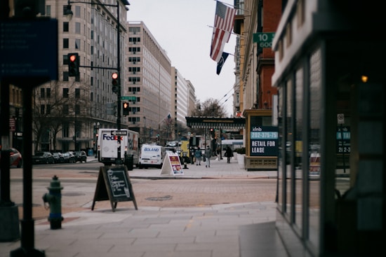 An urban street scene with tall buildings lining both sides. A FedEx truck and other vehicles are on the road. People are walking on the sidewalks, and there are signs advertising retail space for lease. Flags hang from the buildings, and the atmosphere is overcast and calm.