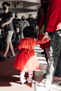 A child wearing a bright red dress with polka-dot sleeves is holding hands with an adult, surrounded by a group of people. The setting appears to be indoors with natural light casting shadows on the floor. The individuals in the background are wearing casual clothing and some have face masks on.