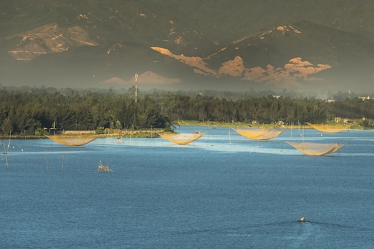A tranquil water scene with large fishing nets suspended over a calm body of blue water, set against a backdrop of lush green hills and forests under a cloudy sky. The early morning or late afternoon light casts a warm glow over the landscape.