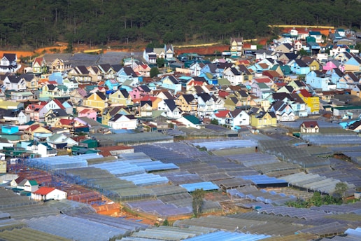 A vibrant town with a cluster of colorful houses, featuring a variety of architectural styles and hues. In the foreground are numerous greenhouses, with a forested area in the background.