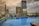 A rooftop infinity pool overlooks a cityscape with several modern skyscrapers under a cloudy sky. The pool is surrounded by a glass railing and a wooden deck with lounge chairs. The buildings reflect the warm hues of a setting or rising sun, creating a serene and luxurious atmosphere.