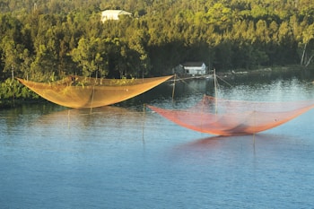 Large fishing nets are suspended over a calm blue lake, with lush green trees and a small building situated on the far shore in the background.