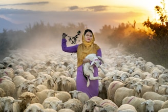 A woman wearing a yellow headscarf and a purple dress stands amidst a large flock of sheep. She is holding a lamb in her arms and has a branch with leaves in her other hand. The scene is set during a sunset, with warm light illuminating the landscape and creating a soft, hazy atmosphere. Bushes and an open field serve as the background.