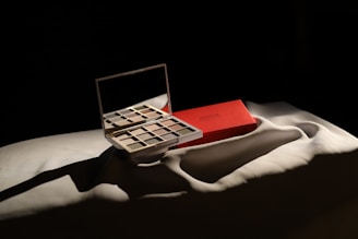 An open makeup palette with a mirror reflects light, revealing a variety of neutral and earthy eyeshadow shades. The palette is placed on a folded white fabric beside a closed red makeup box, which is also partially visible. The scene is dramatically lit, creating deep shadows and highlighting the textures of the fabric and objects.
