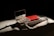 An open makeup palette with a mirror reflects light, revealing a variety of neutral and earthy eyeshadow shades. The palette is placed on a folded white fabric beside a closed red makeup box, which is also partially visible. The scene is dramatically lit, creating deep shadows and highlighting the textures of the fabric and objects.