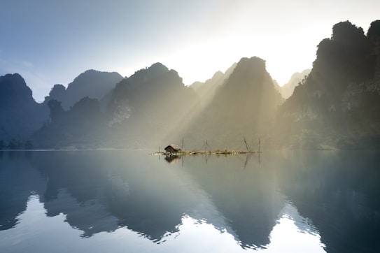 A serene lake reflects towering mountains under a gentle sky. Sunlight filters through the peaks, illuminating a small, rustic hut with a thatched roof surrounded by wooden structures. The calm water mirrors the silhouettes of the landscape, enhancing the tranquil atmosphere.