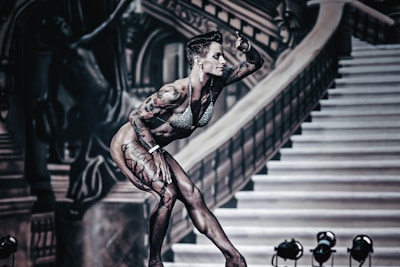 A muscular person with tattoos, wearing a bikini, poses on stage. The background features ornate architectural elements and a staircase with dramatic lighting.
