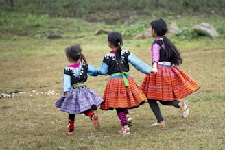 Three children wearing colorful, traditional attire walk hand in hand across a grassy field. Their dresses feature vibrant patterns and they are adorned with decorative elements. The children have long hair, and they appear joyful as they move together.