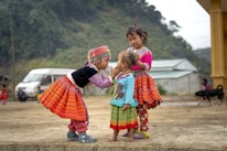 Three young children are dressed in vibrant, traditional clothing. The child on the left reaches out to gently touch the face of the smallest child in the center. The child on the right stands closely, observing with a calm demeanor. The setting appears to be an outdoor, rural area with a background of trees, a van, and a yellow building. There is also a dog in the background.