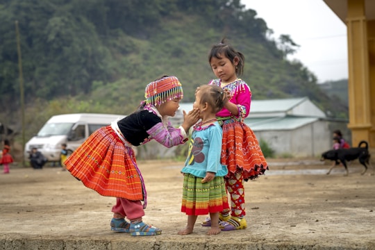 Three young children are dressed in vibrant, traditional clothing. The child on the left reaches out to gently touch the face of the smallest child in the center. The child on the right stands closely, observing with a calm demeanor. The setting appears to be an outdoor, rural area with a background of trees, a van, and a yellow building. There is also a dog in the background.