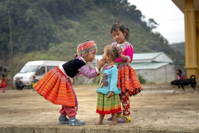 Three young children are dressed in vibrant, traditional clothing. The child on the left reaches out to gently touch the face of the smallest child in the center. The child on the right stands closely, observing with a calm demeanor. The setting appears to be an outdoor, rural area with a background of trees, a van, and a yellow building. There is also a dog in the background.