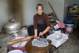 A woman is making rice paper in a simple kitchen setting. She is seated beside a low table covered with rice paper sheets and various utensils. The room is cluttered with cooking equipment, plastic containers, and cloth, suggesting a handmade process.