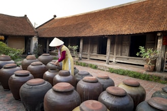 A person wearing traditional clothing and a conical hat is interacting with large ceramic jars placed in a courtyard. The setting features a thatched-roof building in the background, which appears to be a traditional structure with wooden doors and walls. Some greenery is present, including potted plants on the side.