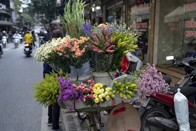 A vibrant assortment of flowers in various colors, including pink, yellow, white, and purple, are arranged in buckets on a bicycle. The bicycle is parked on a busy street, with motorcycles and pedestrians visible in the background. A conical hat rests on the handlebars, adding a cultural touch.