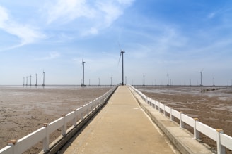 A long concrete pathway leads into the distance, flanked by a white fence on each side. Several large wind turbines are visible in the distance, standing tall in a barren, muddy landscape under a clear blue sky.