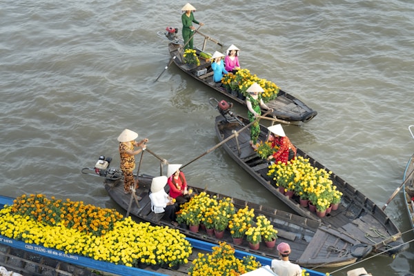 Several wooden boats are on a river, each carrying people wearing traditional conical hats and vibrant clothing. The boats are filled with pots of yellow flowers. The scene appears lively and traditional, showcasing local culture and commerce.