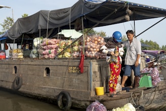 A wooden boat loaded with bags of fresh produce, including onions, potatoes, and other vegetables, floats on the water. Two people are on the boat, one wearing a blue helmet and colorful pants, engaged in conversation. The boat is covered with a tarp, providing some shade for the goods.