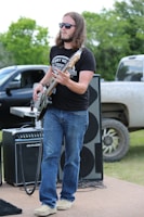 A man with long hair wearing sunglasses plays a bass guitar outdoors. He is dressed in a black t-shirt and blue jeans, and stands in front of large speakers and an amplifier. A black pickup truck is partially visible in the background, with green trees providing a natural backdrop.