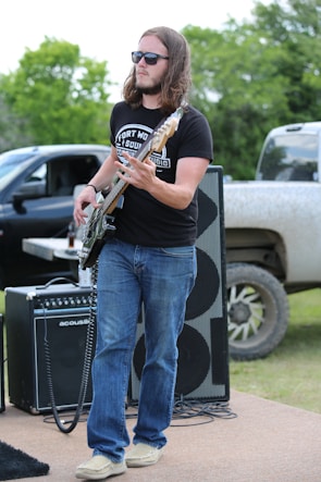 A man with long hair wearing sunglasses plays a bass guitar outdoors. He is dressed in a black t-shirt and blue jeans, and stands in front of large speakers and an amplifier. A black pickup truck is partially visible in the background, with green trees providing a natural backdrop.