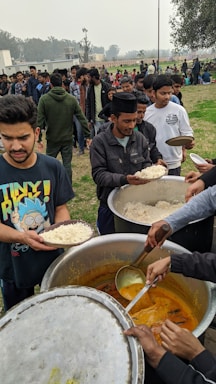 A group of people are standing in line outdoors, receiving food from large pots. Several individuals are holding plates filled with rice while others are serving a yellow curry-like dish. The setting appears to be a casual gathering in a field with trees and buildings in the background.