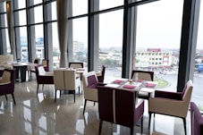 A modern, elegant restaurant setting with large floor-to-ceiling windows that offer a view of an urban landscape. The interior features a polished wooden floor, contemporary dining tables, and chairs with a mix of white and deep purple upholstery. Each table is neatly set with red napkins and cutlery.