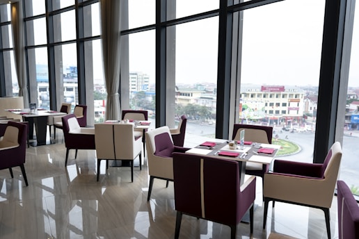 A modern, elegant restaurant setting with large floor-to-ceiling windows that offer a view of an urban landscape. The interior features a polished wooden floor, contemporary dining tables, and chairs with a mix of white and deep purple upholstery. Each table is neatly set with red napkins and cutlery.