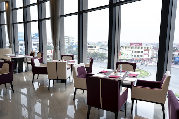 A modern, elegant restaurant setting with large floor-to-ceiling windows that offer a view of an urban landscape. The interior features a polished wooden floor, contemporary dining tables, and chairs with a mix of white and deep purple upholstery. Each table is neatly set with red napkins and cutlery.
