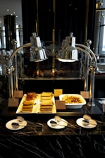 An assortment of breakfast foods displayed under polished silver heat lamps. The foods include stacks of cooked sausages, strips of bacon, and golden-brown potato wedges in a white tray. There are small name tags in front of each type of food. In the foreground, three white plates are set with small spoons and forks on a dark marble counter.