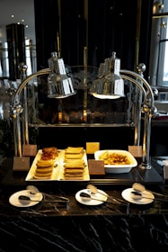 An assortment of breakfast foods displayed under polished silver heat lamps. The foods include stacks of cooked sausages, strips of bacon, and golden-brown potato wedges in a white tray. There are small name tags in front of each type of food. In the foreground, three white plates are set with small spoons and forks on a dark marble counter.