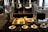 A buffet setup features a variety of breakfast items displayed under shiny metal food warmers. On the countertop, there are trays filled with sausages, bacon, and a bowl of golden potato wedges. Small labels identify each dish. In front, plates with spoons are neatly arranged beside a bowl of red sauce. The setting is elegant, with polished surfaces and ample natural light entering through large windows.