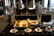 A buffet setup features a variety of breakfast items displayed under shiny metal food warmers. On the countertop, there are trays filled with sausages, bacon, and a bowl of golden potato wedges. Small labels identify each dish. In front, plates with spoons are neatly arranged beside a bowl of red sauce. The setting is elegant, with polished surfaces and ample natural light entering through large windows.