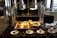 A buffet setup features a variety of breakfast items displayed under shiny metal food warmers. On the countertop, there are trays filled with sausages, bacon, and a bowl of golden potato wedges. Small labels identify each dish. In front, plates with spoons are neatly arranged beside a bowl of red sauce. The setting is elegant, with polished surfaces and ample natural light entering through large windows.
