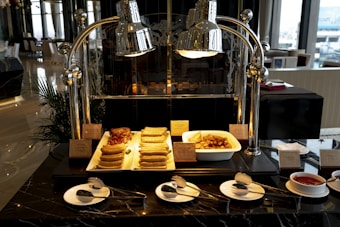 A buffet setup features a variety of breakfast items displayed under shiny metal food warmers. On the countertop, there are trays filled with sausages, bacon, and a bowl of golden potato wedges. Small labels identify each dish. In front, plates with spoons are neatly arranged beside a bowl of red sauce. The setting is elegant, with polished surfaces and ample natural light entering through large windows.