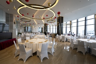 A spacious, elegantly decorated dining area with round tables covered with white cloths, surrounded by white chairs. The ceiling features large circular light fixtures and hanging red and gold ornaments. Large windows provide ample natural light, enhancing the modern and festive atmosphere.