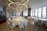 A spacious, elegantly decorated dining area with round tables covered with white cloths, surrounded by white chairs. The ceiling features large circular light fixtures and hanging red and gold ornaments. Large windows provide ample natural light, enhancing the modern and festive atmosphere.