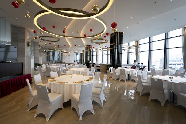 A spacious, elegantly decorated dining area with round tables covered with white cloths, surrounded by white chairs. The ceiling features large circular light fixtures and hanging red and gold ornaments. Large windows provide ample natural light, enhancing the modern and festive atmosphere.