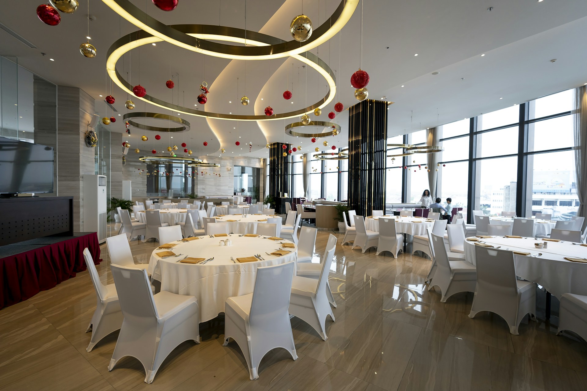 A spacious, elegantly decorated dining area with round tables covered with white cloths, surrounded by white chairs. The ceiling features large circular light fixtures and hanging red and gold ornaments. Large windows provide ample natural light, enhancing the modern and festive atmosphere.