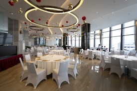 A spacious, elegantly decorated dining area with round tables covered with white cloths, surrounded by white chairs. The ceiling features large circular light fixtures and hanging red and gold ornaments. Large windows provide ample natural light, enhancing the modern and festive atmosphere.