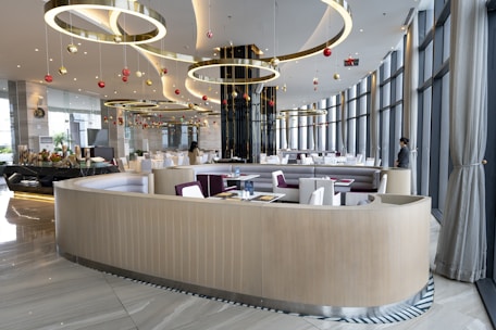 A modern, elegantly designed restaurant interior featuring curvilinear seating arrangements, large floor-to-ceiling windows with curtains, and hanging golden rings adorned with red and gold ornaments. The space is well-lit with soft overhead lighting, and there are a few people present.