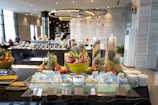 A luxurious buffet setup featuring tropical fruits such as dragon fruit and pineapple arranged in large decorative bowls. Breakfast items, including various cereals and milk cartons, are neatly displayed around a glass ice platter. The background reveals a modern dining area with geometric light fixtures, tables filled with more food items, and staff attending the setup.