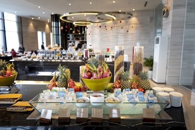 A luxurious buffet setup featuring tropical fruits such as dragon fruit and pineapple arranged in large decorative bowls. Breakfast items, including various cereals and milk cartons, are neatly displayed around a glass ice platter. The background reveals a modern dining area with geometric light fixtures, tables filled with more food items, and staff attending the setup.