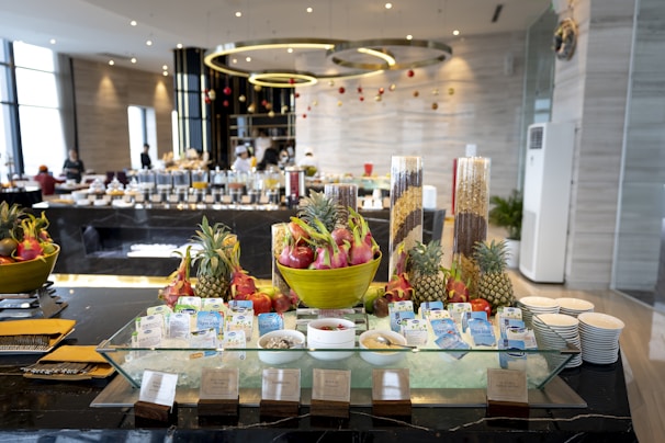 A luxurious buffet setup featuring tropical fruits such as dragon fruit and pineapple arranged in large decorative bowls. Breakfast items, including various cereals and milk cartons, are neatly displayed around a glass ice platter. The background reveals a modern dining area with geometric light fixtures, tables filled with more food items, and staff attending the setup.